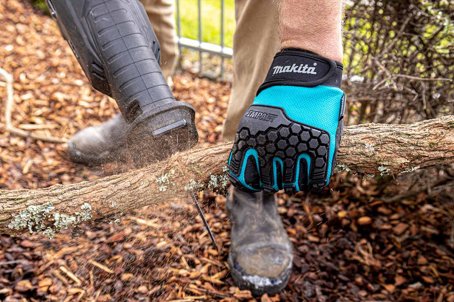 A close-up action shot of a person using a chainsaw to cut down a fallen tree branch in a mulched outdoor area. The person is wearing a black and teal Makita work glove on their hand, along with black boots and shoes.