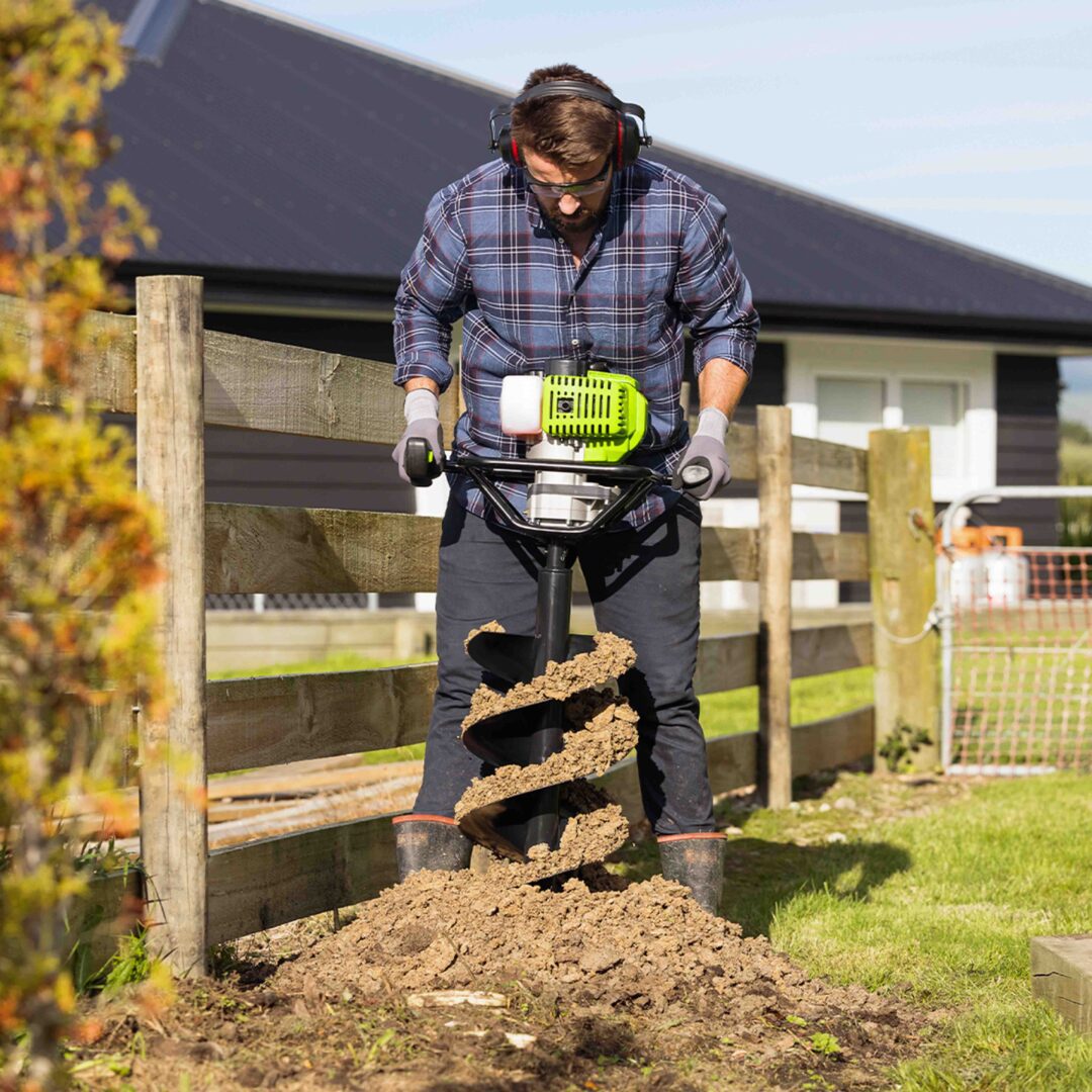 Person using a soil auger in a garden setting with a house in the background