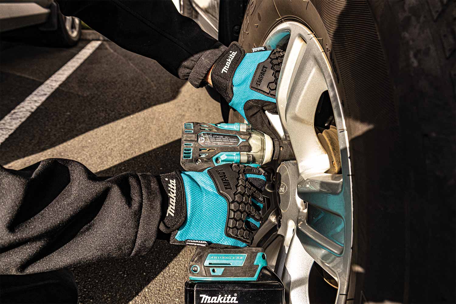 A person wearing teal and black Makita gloves uses a cordless impact wrench to remove a lug from a silver car wheel in an outdoor parking lot. The Makita brand is clearly visible on the gloves, tool, and battery pack.