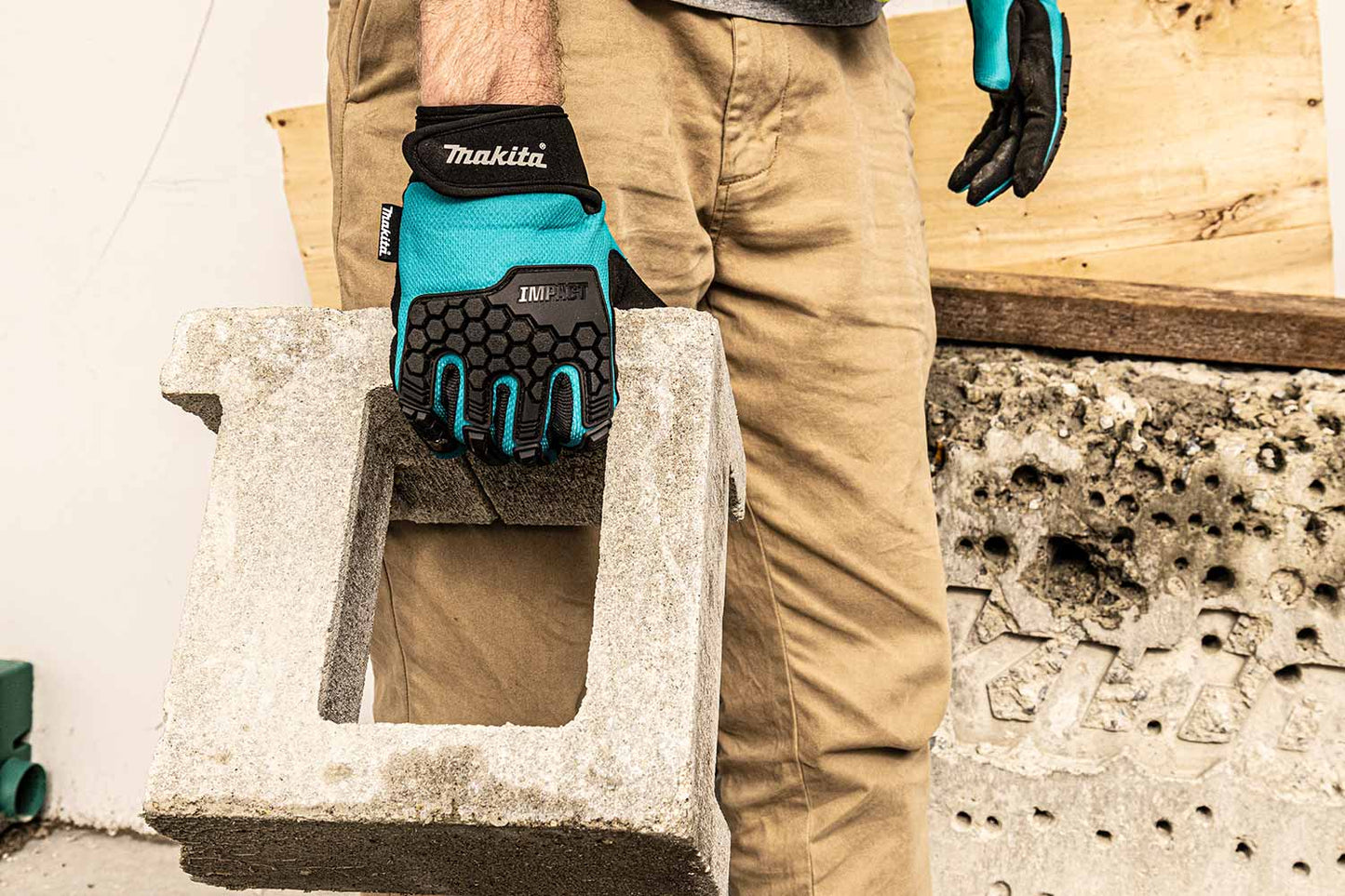 A person wearing tan work pants and teal and black Makita work gloves holds a concrete block in a workshop setting.