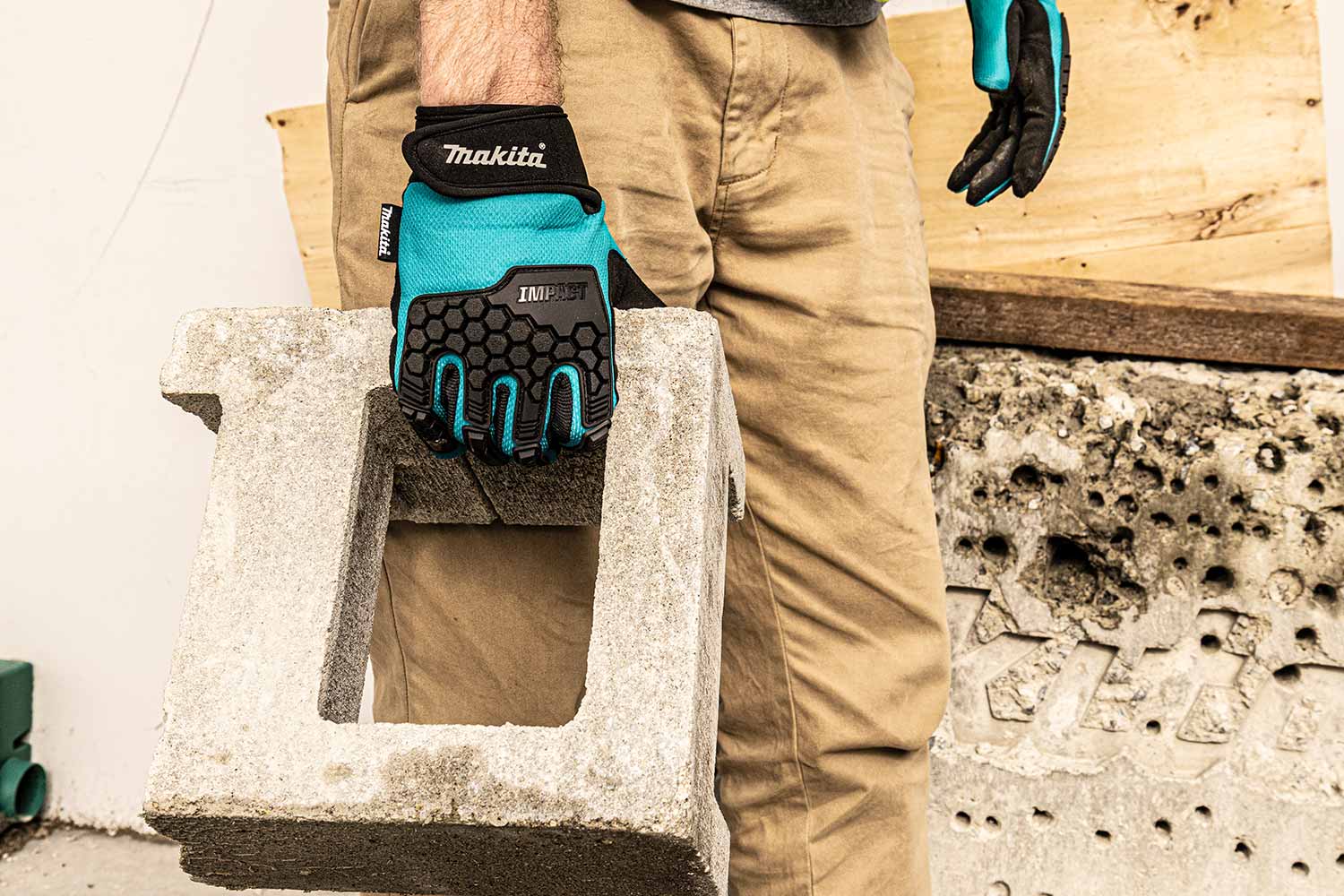 A person wearing tan work pants and teal and black Makita work gloves holds a concrete block in a workshop setting.
