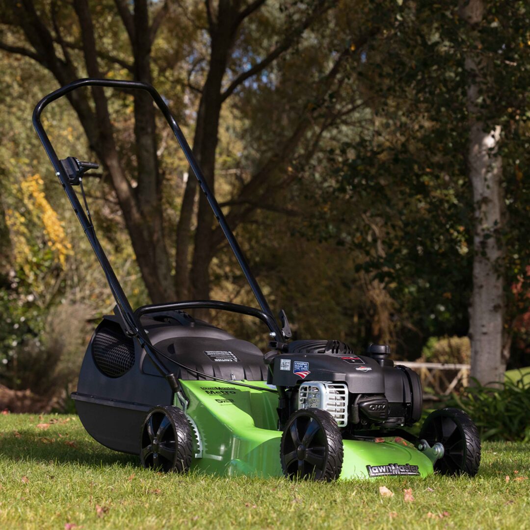 A green and black LawnMaster lawn mower with a black handle is positioned on a grassy lawn with trees in the background.