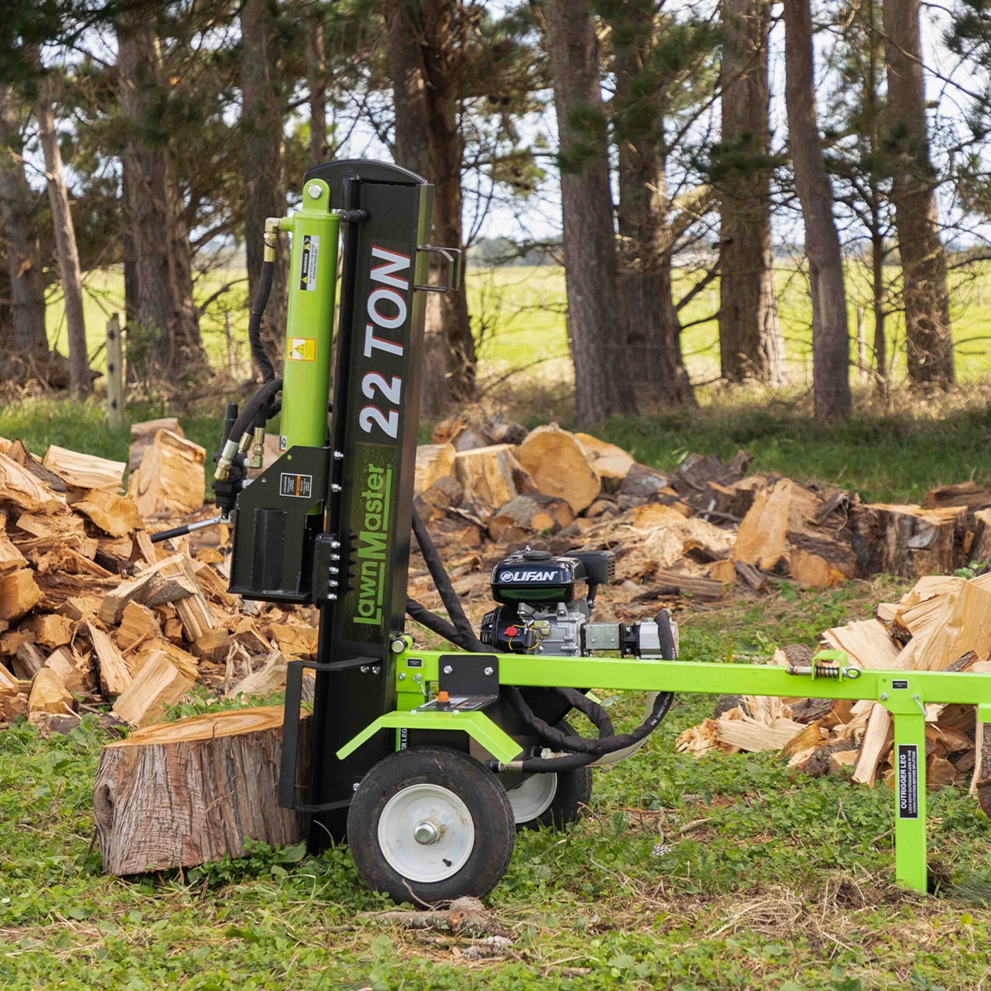 Green and black log splitter in a forest setting with stacked logs around it.