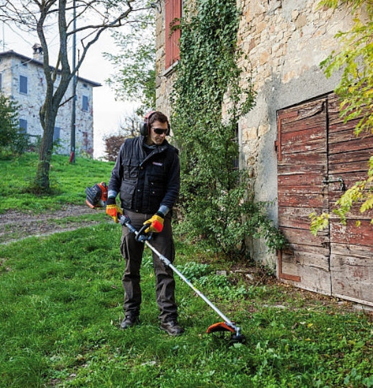 Person using an Oleo-Mac grass trimmer in a garden setting with a rustic building in the background