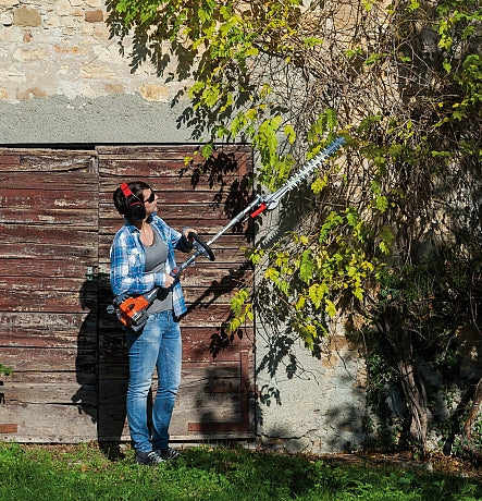 Person using an Oleo-Mac pole hedge trimmer in front of a wooden shed and stone wall.