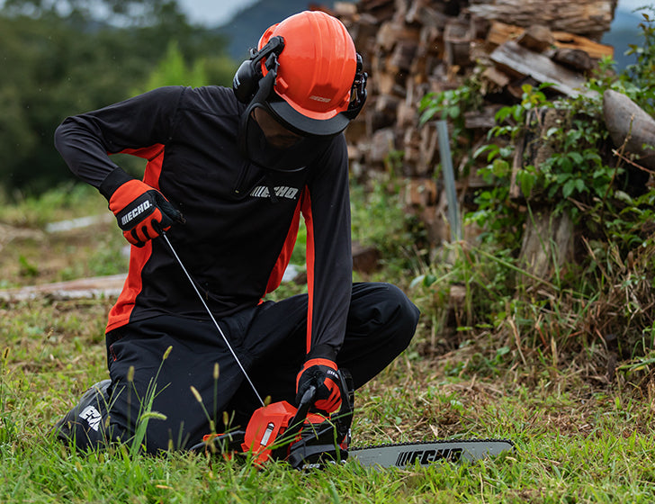 A person with an echo cs-3410 chainsaw, in a black and orange work uniform kneels in a grassy outdoor area, holding a chainsaw with a spark arrestor and wearing a bright orange hard hat and ear protection. A stack of firewood is visible in the background.