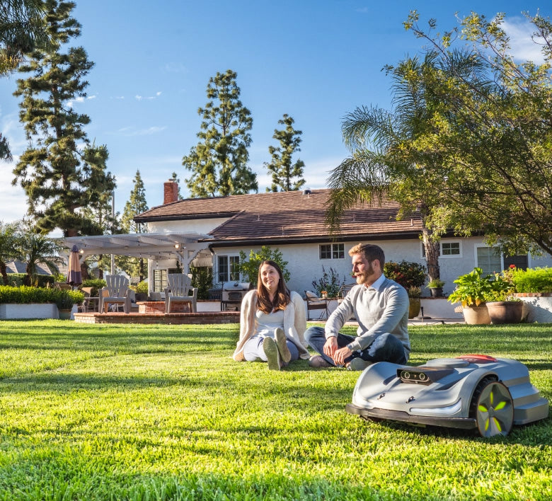 A man and a woman sit on a green lawn in front of a suburban house, with a robotic lawnmower in the foreground. The scene is set in a real outdoor environment with trees, a house, and patio furniture visible.