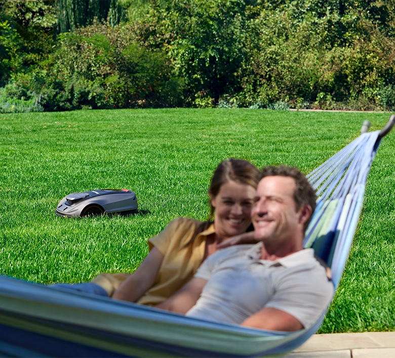 A man and woman relax in a blue and white striped outdoor hammock on a lawn, with a robotic lawnmower visible in the background.